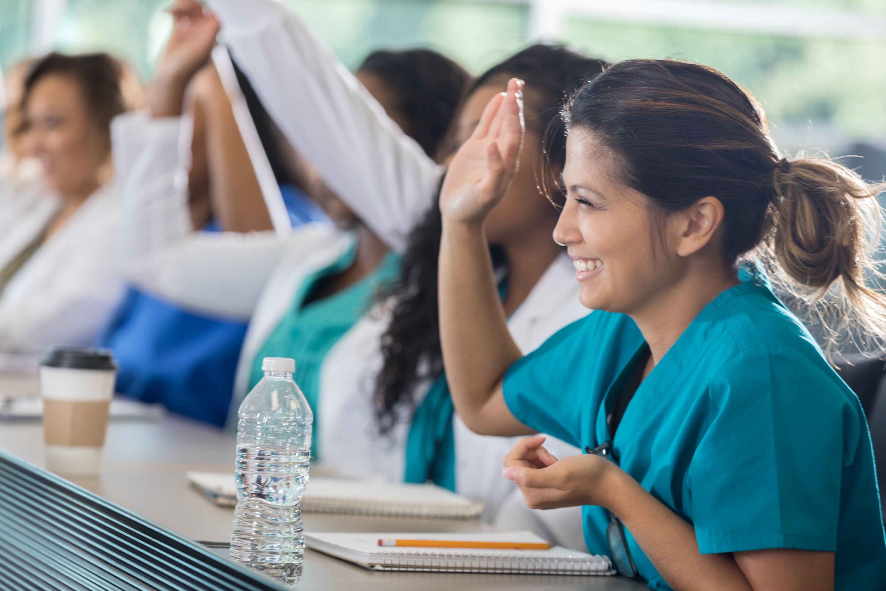 Cheerful Asian female medical student raises her hand to answer a question during class. She is wearing scrubs. A notebook and a bottle of water are on the table in front of her.